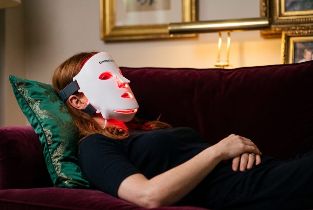 Woman resting on a burgundy velvet sofa wearing the CurrentBody LED Face Mask Series 3