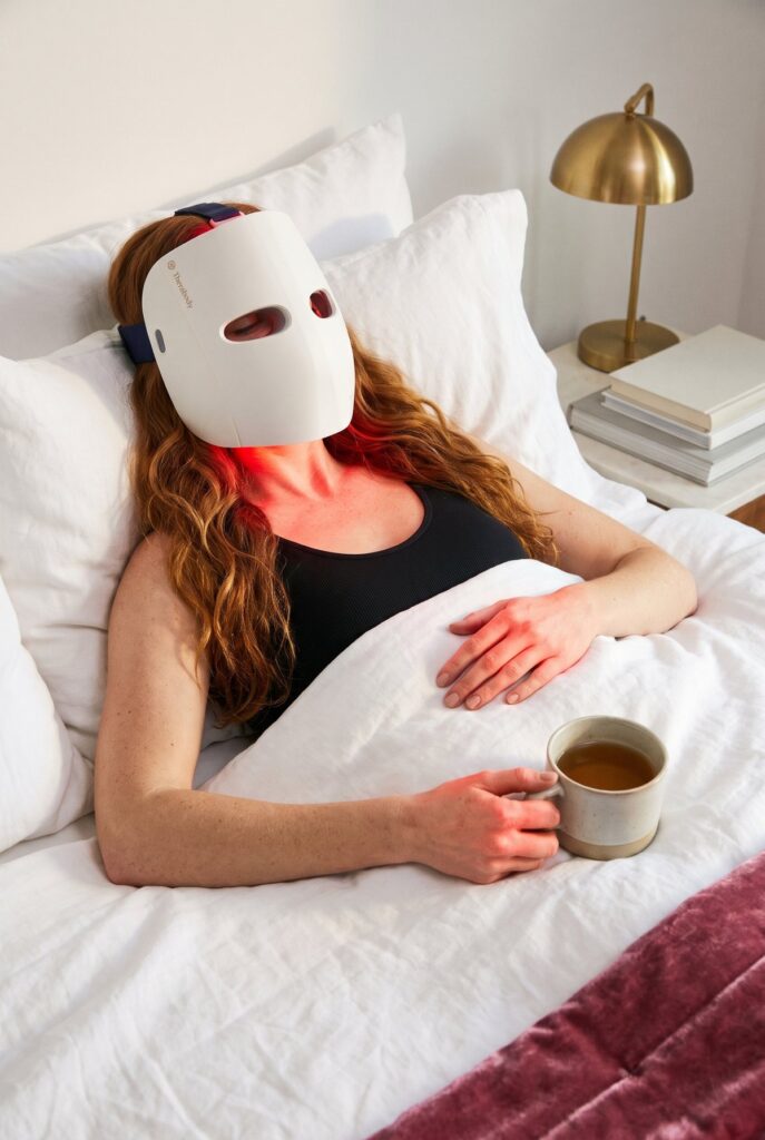 Woman relaxing in bed with a cup of tea while wearing the Therabody TheraFace Mask Glo
